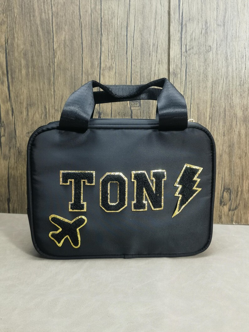 K&ouml;nnte beinhalten: Schwarze Reisetasche mit einem oberen Griff und einem goldenen Rei&szlig;verschluss. Die Tasche zeigt das Wort "TON" in goldenen, umrandeten Buchstaben, zusammen mit einem goldenen Flugzeug und Blitz-Grafik. Die Tasche ist rechteckig und hat einen schwarzen Riemen.
