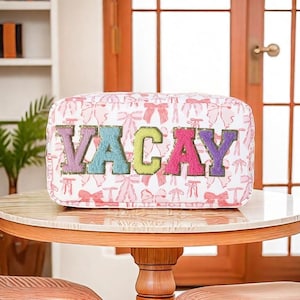 Op de afbeelding: Een rechthoekige make-uptas met het woord "VACAY" in kleurrijke, getextureerde letters. De tas heeft een witte achtergrond met een roze strikpatroon. De tas staat op een ronde tafel.