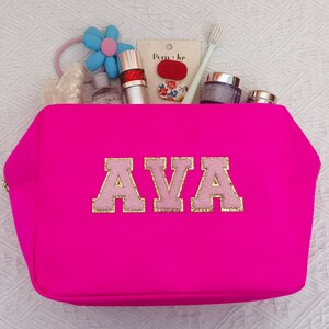 May include: A bright pink zippered pouch with the name "AVA" in pink and gold letters. The pouch is filled with various items, including a toothbrush, hair ties, and cosmetics.