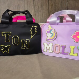 K&ouml;nnte beinhalten: Zwei kleine, rechteckige Taschen mit Griffen. Eine ist schwarz mit goldenen Buchstaben, die "TON" buchstabieren, und einem Blitz- und Flugzeugaufn&auml;her. Die andere ist lavendelfarben mit "MOLLY" in bunten Buchstaben und einem Einhorn- und Blumenaufn&auml;her.