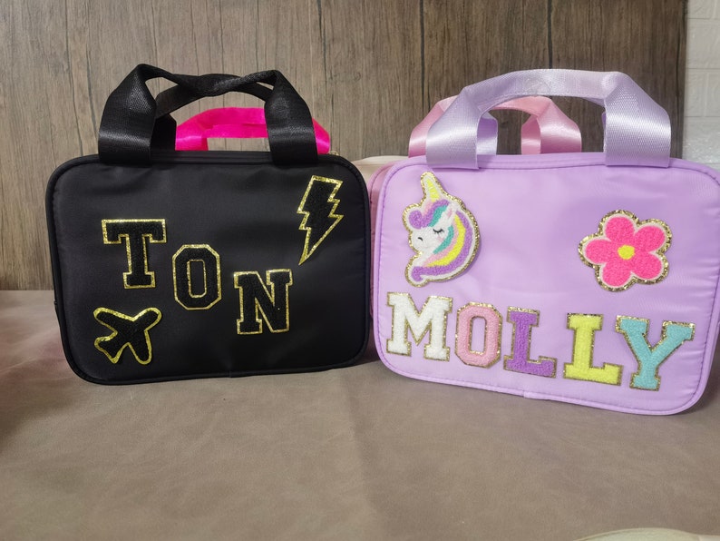 K&ouml;nnte beinhalten: Zwei kleine, rechteckige Taschen mit Griffen. Eine ist schwarz mit goldenen Buchstaben, die "TON" buchstabieren, und einem Blitz- und Flugzeugaufn&auml;her. Die andere ist lavendelfarben mit "MOLLY" in bunten Buchstaben und einem Einhorn- und Blumenaufn&auml;her.