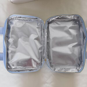 K&ouml;nnte beinhalten: Ge&ouml;ffnete, hellblaue isolierte Lunchtasche mit silbernem Innenraum. Die Tasche hat einen Rei&szlig;verschluss und zwei hellblaue Griffe. Der Innenraum ist mit einem reflektierenden, metallischen Material ausgekleidet.