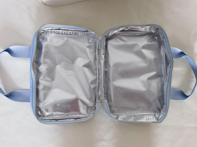 K&ouml;nnte beinhalten: Ge&ouml;ffnete, hellblaue isolierte Lunchtasche mit silbernem Innenraum. Die Tasche hat einen Rei&szlig;verschluss und zwei hellblaue Griffe. Der Innenraum ist mit einem reflektierenden, metallischen Material ausgekleidet.