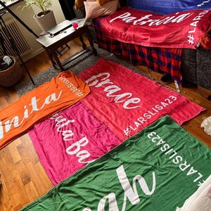 Pu&ograve; includere: Cinque coperte colorate con testo bianco. Le coperte sono disposte su un pavimento di legno. Il testo sulle coperte recita "nita", "uta Ba", "Grace", "Lisa A! Patricia" e "leah".