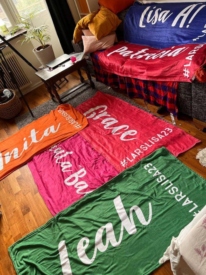 Pu&ograve; includere: Cinque coperte colorate con testo bianco. Le coperte sono disposte su un pavimento di legno. Il testo sulle coperte recita "nita", "uta Ba", "Grace", "Lisa A! Patricia" e "leah".