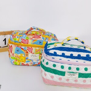 May include: Two quilted cosmetic bags with top handles. One bag is patterned with colorful cartoon characters, hearts, and rainbows. The other bag has a white background with blue stripes and green polka dots. Both bags have a zipper closure and a small tag that reads "today mood feel the warm."