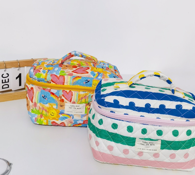 May include: Two quilted cosmetic bags with top handles. One bag is patterned with colorful cartoon characters, hearts, and rainbows. The other bag has a white background with blue stripes and green polka dots. Both bags have a zipper closure and a small tag that reads "today mood feel the warm."