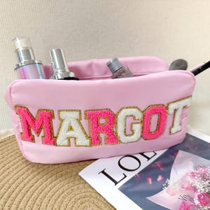 Pode incluir: Uma bolsa de maquiagem rosa com o nome "MARGOT" bordado em letras rosa e brancas com acabamento dourado. A bolsa contém produtos de maquiagem e um pincel. Uma revista e um buquê de rosas cor-de-rosa estão ao fundo.