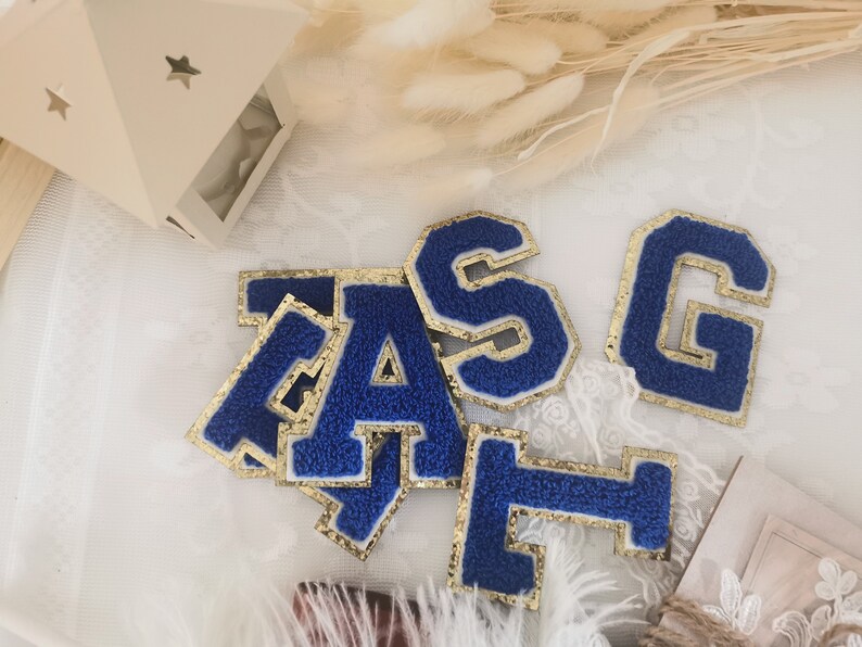 May include: Blue felt letters with gold glitter trim, spelling out the word "TAGS". The letters are on a white surface with a white and gold lace overlay.
