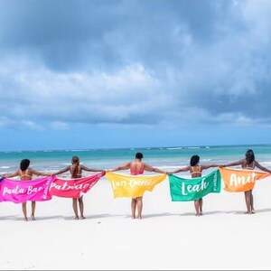 Pu&ograve; includere: Sette persone su una spiaggia che tengono asciugamani colorati con i loro nomi scritti su di essi. Gli asciugamani sono blu, rosa, rosso, giallo, verde, arancione e verde. Le persone sono in fila rivolte verso l'oceano.