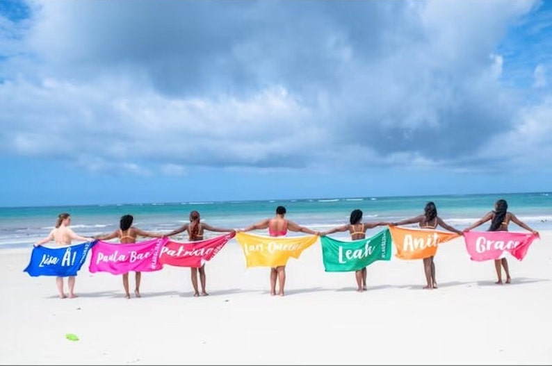 Pu&ograve; includere: Sette persone su una spiaggia che tengono asciugamani colorati con i loro nomi scritti su di essi. Gli asciugamani sono blu, rosa, rosso, giallo, verde, arancione e verde. Le persone sono in fila rivolte verso l'oceano.
