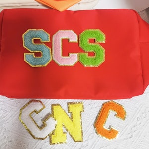 May include: Red fabric pouch with the letters SCS in colorful felt patches. Below the pouch are three more felt patches, spelling out CNC, in white, yellow, and orange.