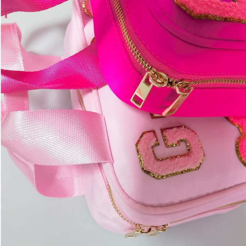 K&ouml;nnte beinhalten: Zwei rosa Rucks&auml;cke, einer hellrosa und einer pink, sind gestapelt. Der hellrosa Rucksack hat einen goldenen Rei&szlig;verschluss und einen flauschigen Buchstabenaufn&auml;her. Der pinkfarbene Rucksack hat einen goldenen Rei&szlig;verschluss. Beide haben rosa Tr&auml;ger.