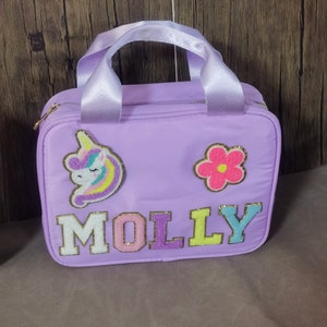 K&ouml;nnte beinhalten: Eine lavendelfarbene Tragetasche mit wei&szlig;em Griff und goldenem Rei&szlig;verschluss. Die Tasche hat einen Einhorn- und Blumenaufn&auml;her und den Namen "MOLLY" in bunten, strukturierten Buchstaben.