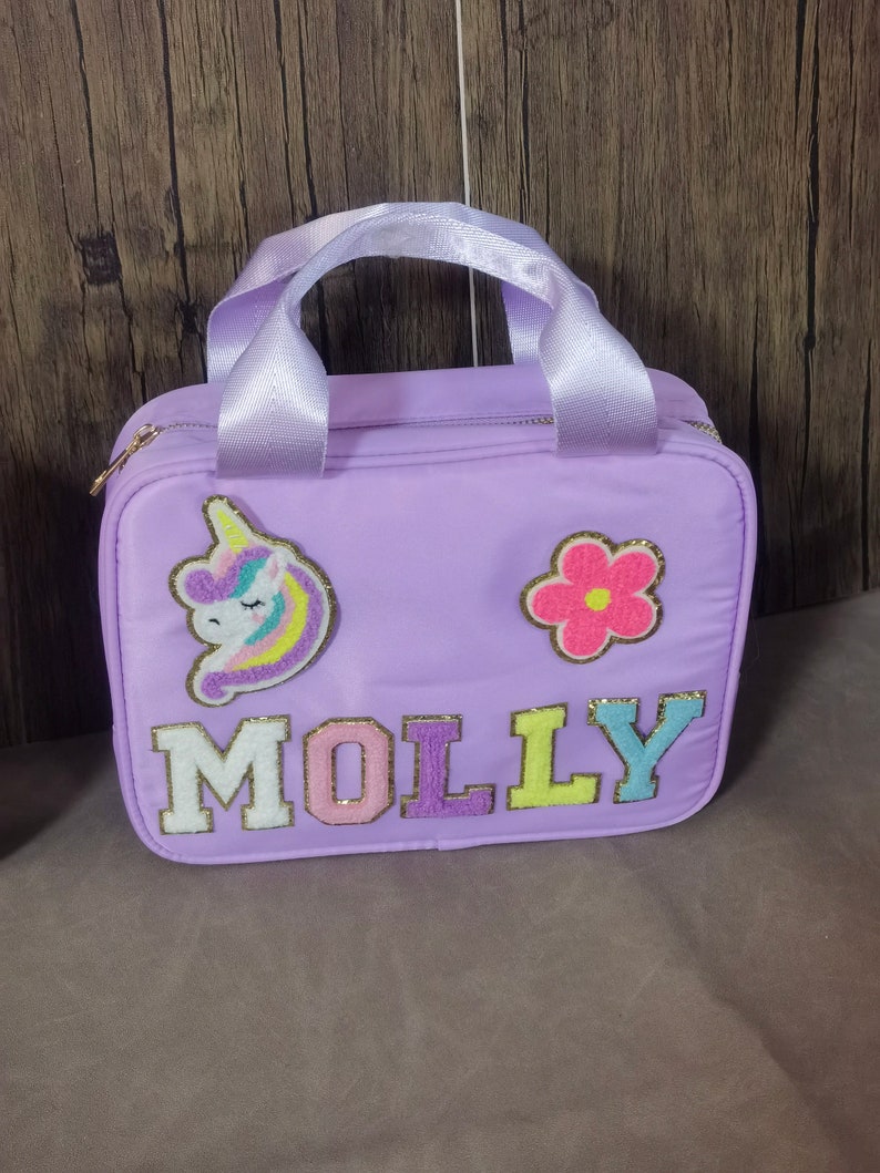 K&ouml;nnte beinhalten: Eine lavendelfarbene Tragetasche mit wei&szlig;em Griff und goldenem Rei&szlig;verschluss. Die Tasche hat einen Einhorn- und Blumenaufn&auml;her und den Namen "MOLLY" in bunten, strukturierten Buchstaben.