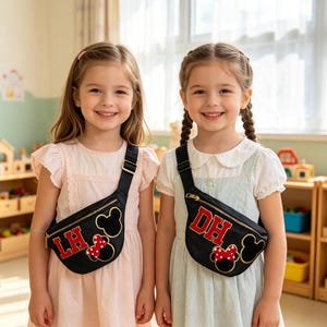 May include: Two black fanny packs featuring gold-outlined Mickey Mouse silhouettes and red-and-white polka dot bows. One bag displays the red letters LH, and the other displays the red letters DH. Each bag has a black adjustable strap and gold zipper.