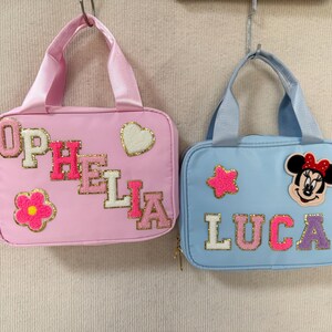 K&ouml;nnte beinhalten: Zwei kleine, rechteckige Handtaschen mit Henkeln h&auml;ngen vor einem neutralen Hintergrund. Eine ist rosa mit dem Namen "OPHELIA" und einer Blumen- und Herzverzierung. Die andere ist hellblau mit "LUCA" und einem Minnie-Mouse-Aufn&auml;her.