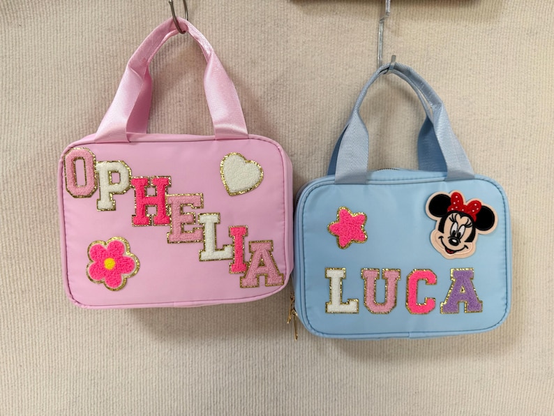 K&ouml;nnte beinhalten: Zwei kleine, rechteckige Handtaschen mit Henkeln h&auml;ngen vor einem neutralen Hintergrund. Eine ist rosa mit dem Namen "OPHELIA" und einer Blumen- und Herzverzierung. Die andere ist hellblau mit "LUCA" und einem Minnie-Mouse-Aufn&auml;her.