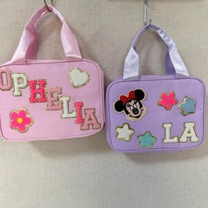 K&ouml;nnte beinhalten: Zwei kleine, rechteckige Handtaschen mit Griffen. Eine ist rosa mit dem Namen "OPHELIA" in bunten Buchstaben und einer Blume und einem Herz. Die andere ist lavendelfarben mit "LA" und einem Minnie-Mouse-Motiv und Sternen.