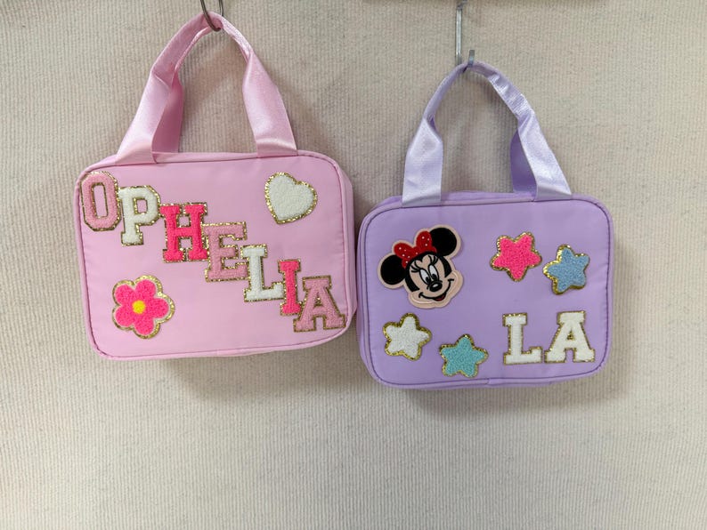 K&ouml;nnte beinhalten: Zwei kleine, rechteckige Handtaschen mit Griffen. Eine ist rosa mit dem Namen "OPHELIA" in bunten Buchstaben und einer Blume und einem Herz. Die andere ist lavendelfarben mit "LA" und einem Minnie-Mouse-Motiv und Sternen.