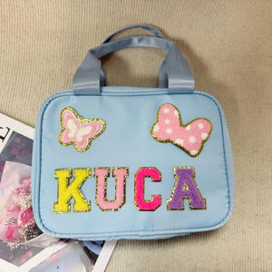 K&ouml;nnte beinhalten: Hellblaue Handtasche mit oberem Griff, verziert mit einem Schmetterling und einer Schleife. Das Wort "KUCA" ist in bunten, glitzernden Buchstaben geschrieben. Im Hintergrund ist eine Zeitschrift mit dem Wort "LOEWE" zu sehen.