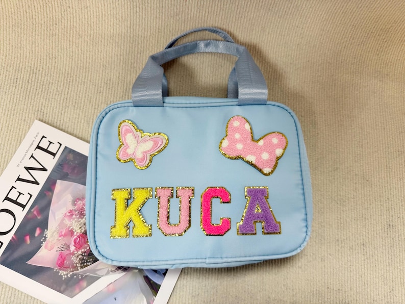 K&ouml;nnte beinhalten: Hellblaue Handtasche mit oberem Griff, verziert mit einem Schmetterling und einer Schleife. Das Wort "KUCA" ist in bunten, glitzernden Buchstaben geschrieben. Im Hintergrund ist eine Zeitschrift mit dem Wort "LOEWE" zu sehen.