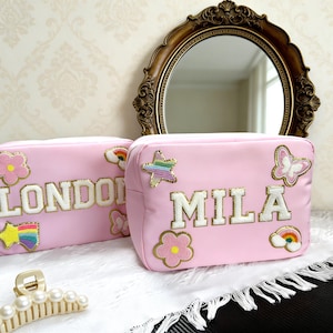 以下が含まれることがあります： 「LONDON」と「MILA」の名前が刺繍されたパッチが付いた2つのピンク色の化粧ポーチ。バッグには金色のジッパーが付いており、花、星、虹、蝶で飾られています。 金色の真珠のヘアクリップが前景にあります。