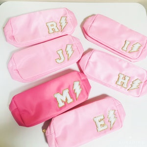 Puede incluir: Seis estuches de satén rosa con cremallera, con bordado de rayo dorado y letras. Las letras son R, J, H, M y E.