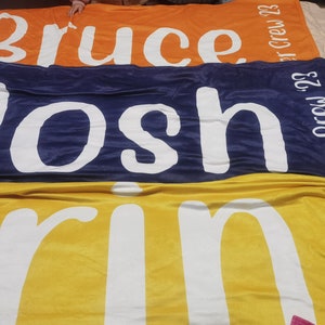 Pu&ograve; includere: Tre coperte in pile impilate una sopra l'altra. La coperta superiore &egrave; arancione con il nome "Bruce" in lettere bianche e il testo "Crew '23". La coperta centrale &egrave; blu scuro con il nome "Josh" in lettere bianche e il testo "Summer Crew '23". La coperta inferiore &egrave; gialla con il nome "Erin" in lettere bianche e il testo "Fall Crew '23".