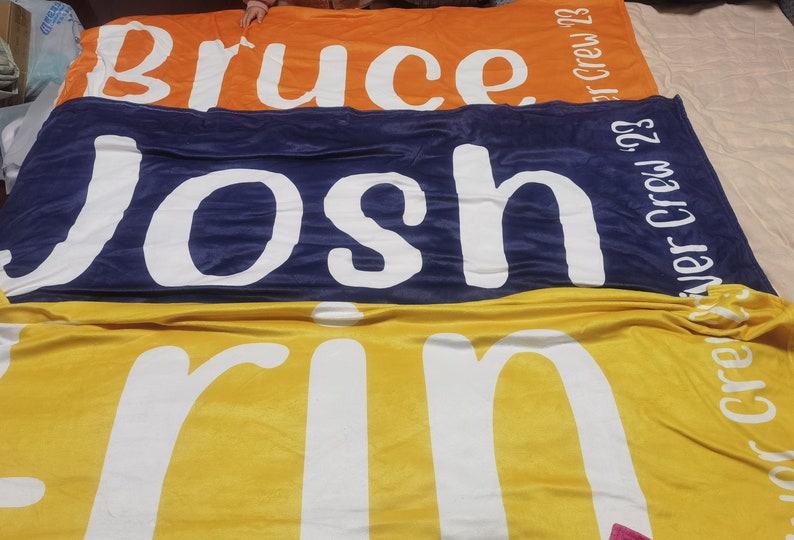 Pu&ograve; includere: Tre coperte in pile impilate una sopra l'altra. La coperta superiore &egrave; arancione con il nome "Bruce" in lettere bianche e il testo "Crew '23". La coperta centrale &egrave; blu scuro con il nome "Josh" in lettere bianche e il testo "Summer Crew '23". La coperta inferiore &egrave; gialla con il nome "Erin" in lettere bianche e il testo "Fall Crew '23".