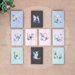May include: A selection of notebooks, each with a butterfly and initial design. The notebooks display different letters, like "R", "A", and "G", accompanied by a vibrant butterfly illustration. The notebooks come in various colors, including light blue, black, and pink.