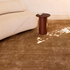 May include: A brown rug with a textured surface, placed in a room with a beige sofa and a small, dark brown side table. A white bowl sits on the table. The room has a neutral color palette and a minimalist design.