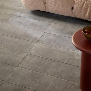 May include: A close-up of a gray rug with a textured surface. The rug is rectangular and has horizontal lines. A light pink, textured cushion is in the background. A small, round, red table with a bowl is in the foreground.