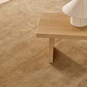 May include: A light brown, rectangular rug with a textured surface. A light wood table with a single leg is on the right, with a white paper lantern on top. The rug and table create a warm, inviting atmosphere.
