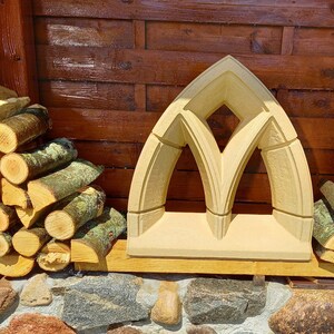 Könnte beinhalten: Ein Steinbogen mit einer spitzen Oberseite und einem rautenförmigen Fenster in der Mitte. Der Bogen ist hellbeige und steht auf einer Mauer aus gestapelten Steinen.