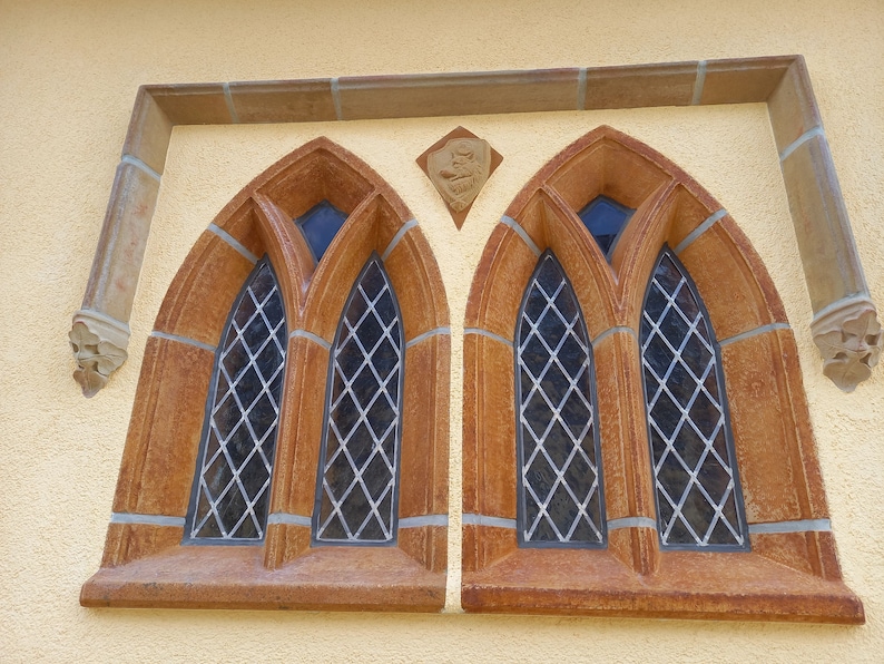 May include: Two arched windows with diamond-shaped glass panes, framed in brown stone, set into a yellow wall.