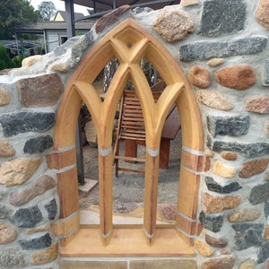 May include: A stone wall with a large, arched window made of brown brick. The window is divided into three sections by thin, vertical strips of brick. The wall is made of rough, uneven stones in shades of gray, brown, and tan.