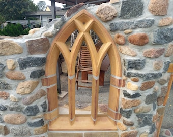 May include: A stone wall with a large, arched window made of brown brick. The window is divided into three sections by thin, vertical strips of brick. The wall is made of rough, uneven stones in shades of grey, brown, and tan.