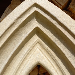 May include: A beige stone architectural element with a pointed arch shape featuring recessed, stepped molding along the edges. The interior of the arch reveals a collection of stacked, light-colored wooden logs with visible grain.