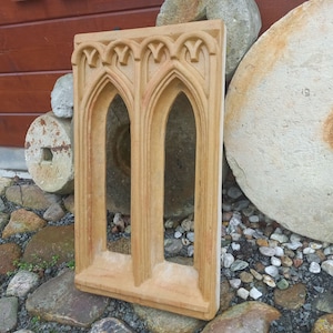 May include: A rectangular tan stone window frame featuring two arched openings with pointed tops and decorative molding along the upper edge. The structure is made of a light brown, textured material with a smooth finish on the front surface.