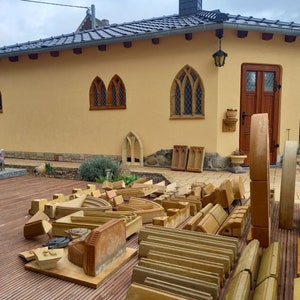 May include: A collection of tan sandstone architectural elements, including curved arches, rectangular blocks, and carved moldings, arranged on a wooden deck in front of a yellow building with a dark tiled roof and wooden door.
