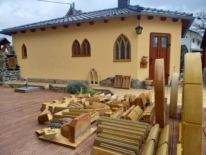May include: A collection of tan sandstone architectural elements, including curved arches, rectangular blocks, and carved moldings, arranged on a wooden deck in front of a yellow building with a dark tiled roof and wooden door.