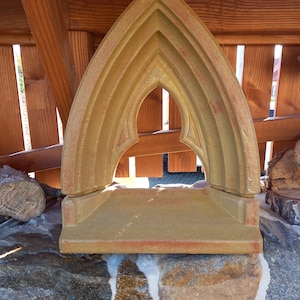 Könnte beinhalten: Ein dekoratives, hellbraunes, gewölbtes Steinstück mit einer spitzen Oberseite. Der Stein hat ein geschnitztes, kompliziertes Design mit mehreren Bögen. Der Stein liegt auf einer Mauer aus gestapelten Steinen.