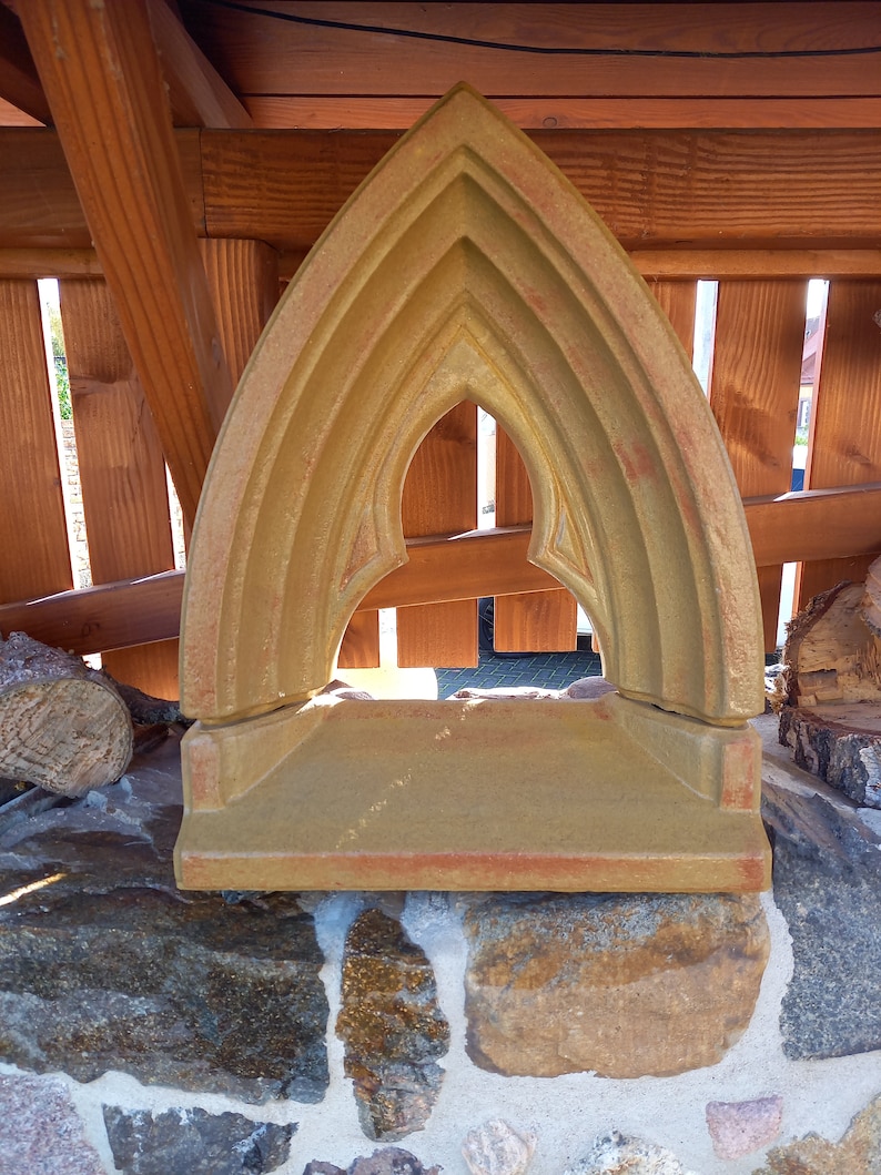 May include: A tan-colored stone arch with a pointed top and a flat base. The structure features a recessed, multi-layered frame design with a central opening, showcasing a weathered, earthy texture with subtle reddish-brown color variations.