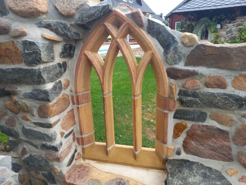 May include: A stone wall with a decorative archway made of brown and red brick. The archway has a pointed top and three vertical panels. The archway is set into the wall and looks out onto a grassy area.