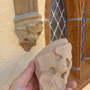 May include: A close-up of a hand holding a light brown stone carving of three leaves. The carving is detailed and intricate, with a textured surface. The carving is similar to one already installed on a wall.