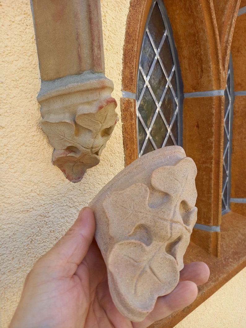 May include: A close-up of a hand holding a light brown stone carving of three leaves. The carving is detailed and intricate, with a textured surface. The carving is similar to one already installed on a wall.