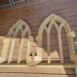 May include: Two unfinished wooden arched window frames, each with three arched openings, are laid out on a wooden deck. The frames are made of multiple pieces of wood, and the arches are formed by overlapping pieces of wood.