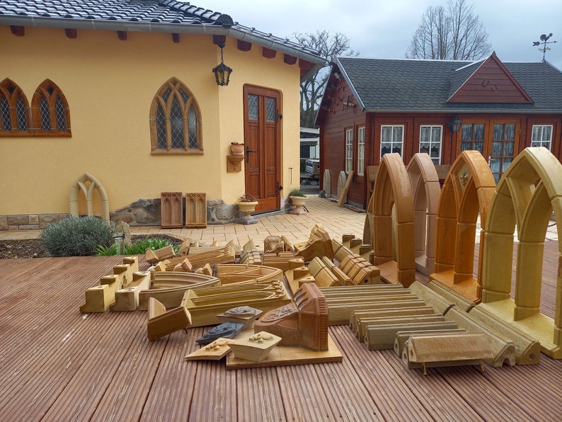 May include: A collection of unfinished, light brown, wooden window frames with arched tops. The frames are arranged on a wooden deck in front of a yellow building with a brown door.