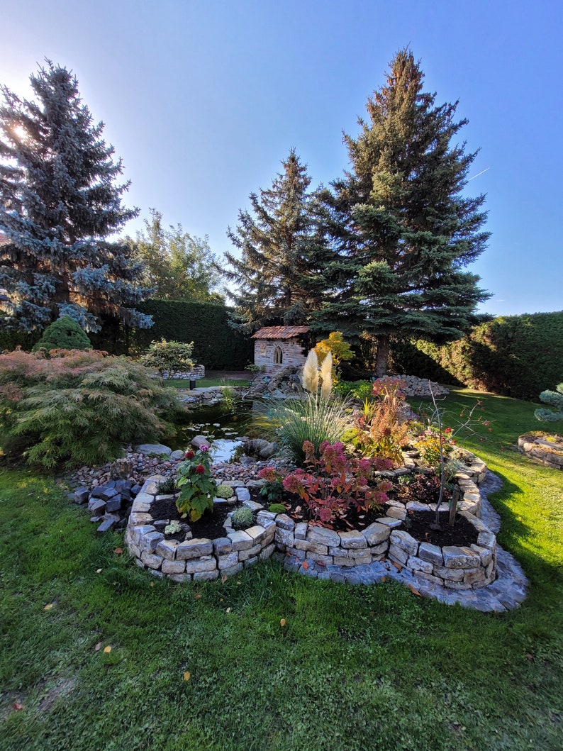 May include: A garden landscape featuring a stone-walled pond with tiered flower beds, various shrubs, and tall evergreen trees. A small stone structure with a tiled roof sits near the water, surrounded by green grass and rocky borders.